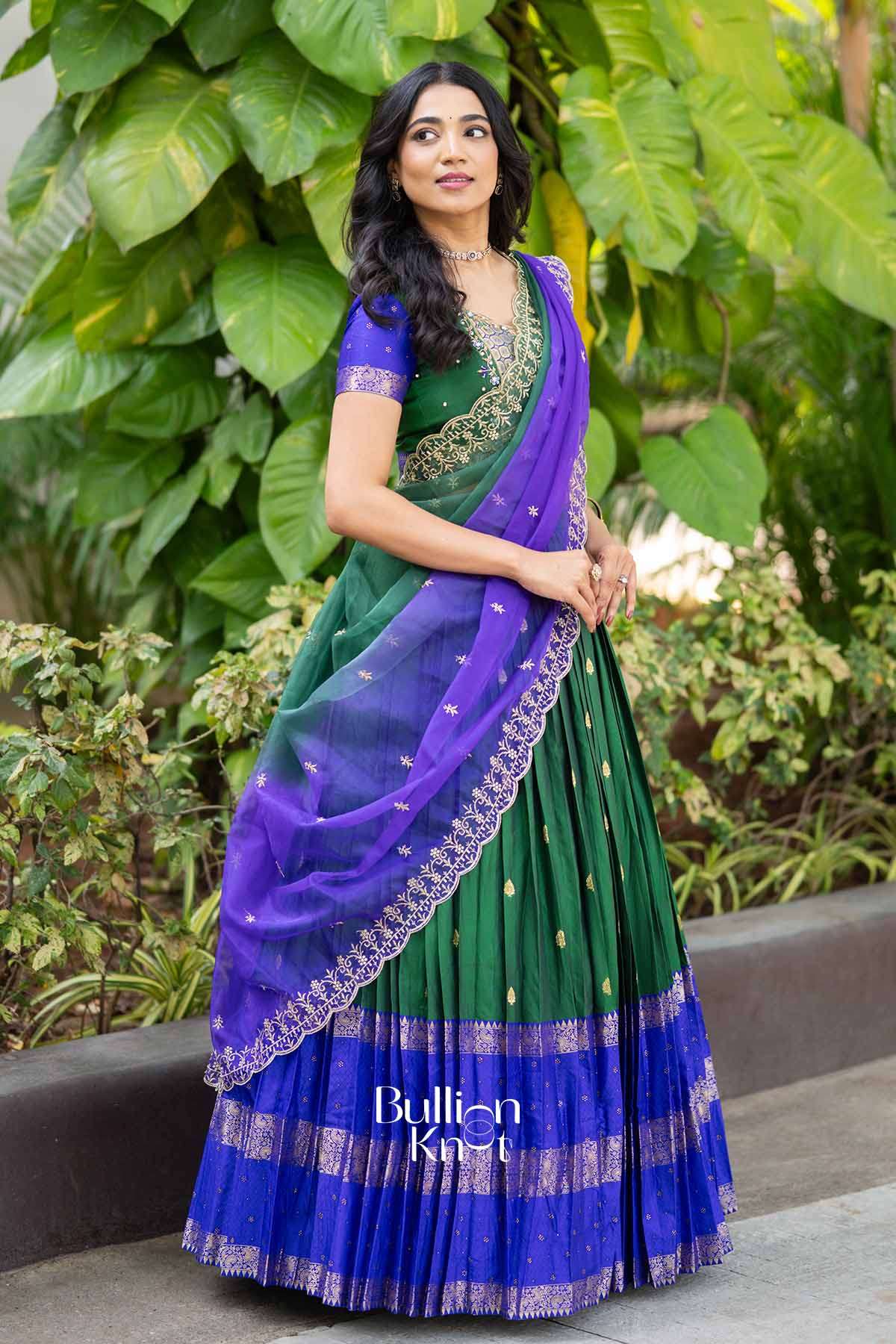 Elegant woman in a green and blue traditional lehenga with intricate gold embroidery, paired with a purple dupatta, standing outdoors among lush green leaves