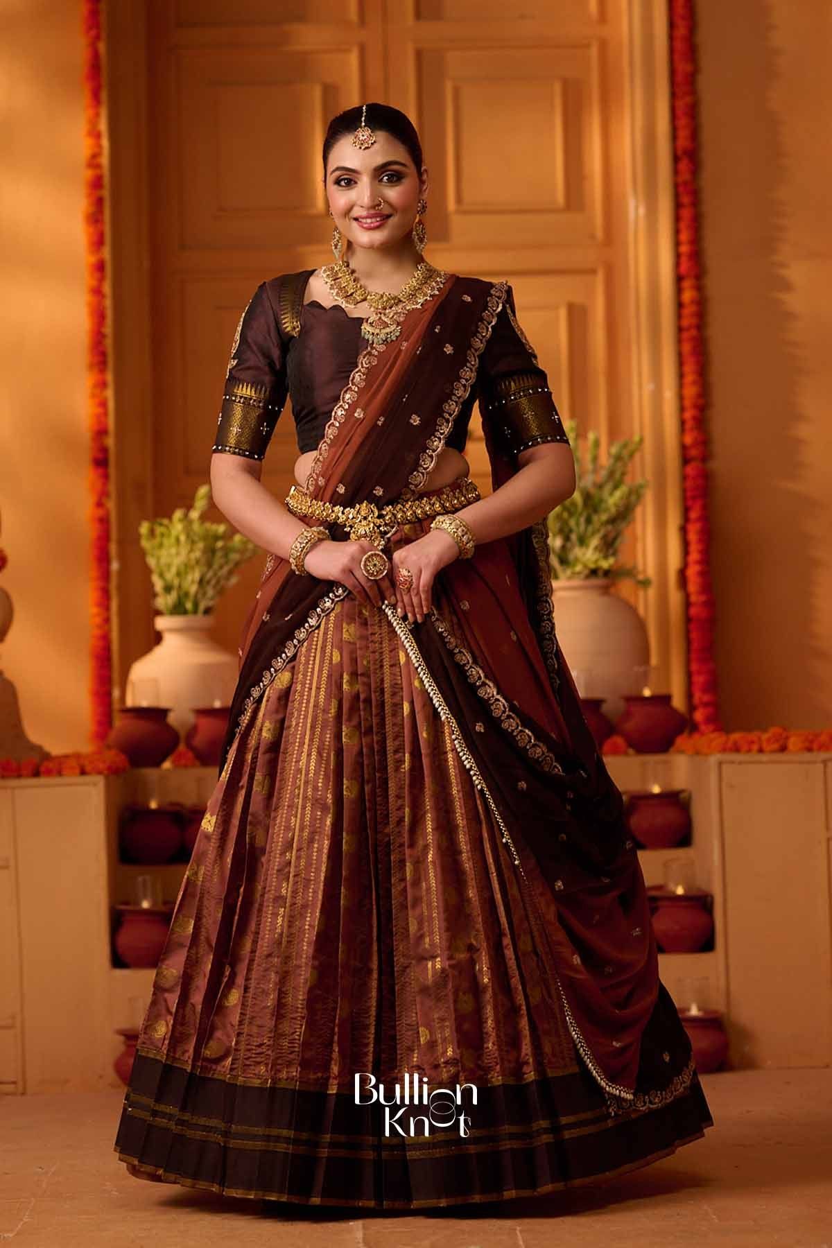 Elegant woman in dark brown and gold lehenga with intricate embroidery, wearing heavy gold jewelry, standing in decorated indoor setting