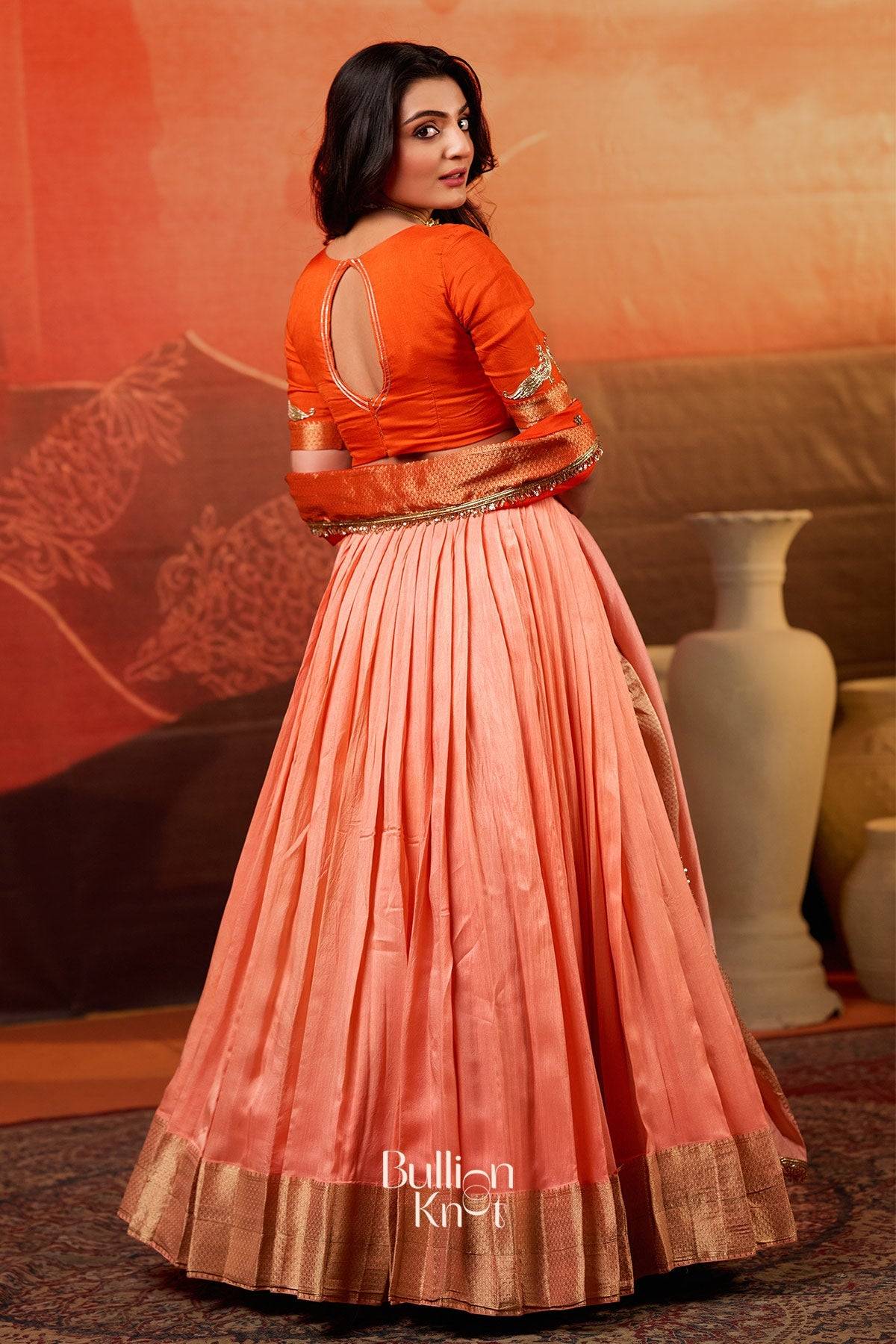 Back view of Kaavya Dola Lehenga Set in Orange at bullionknot, displaying pleated skirt and keyhole blouse design with golden border.