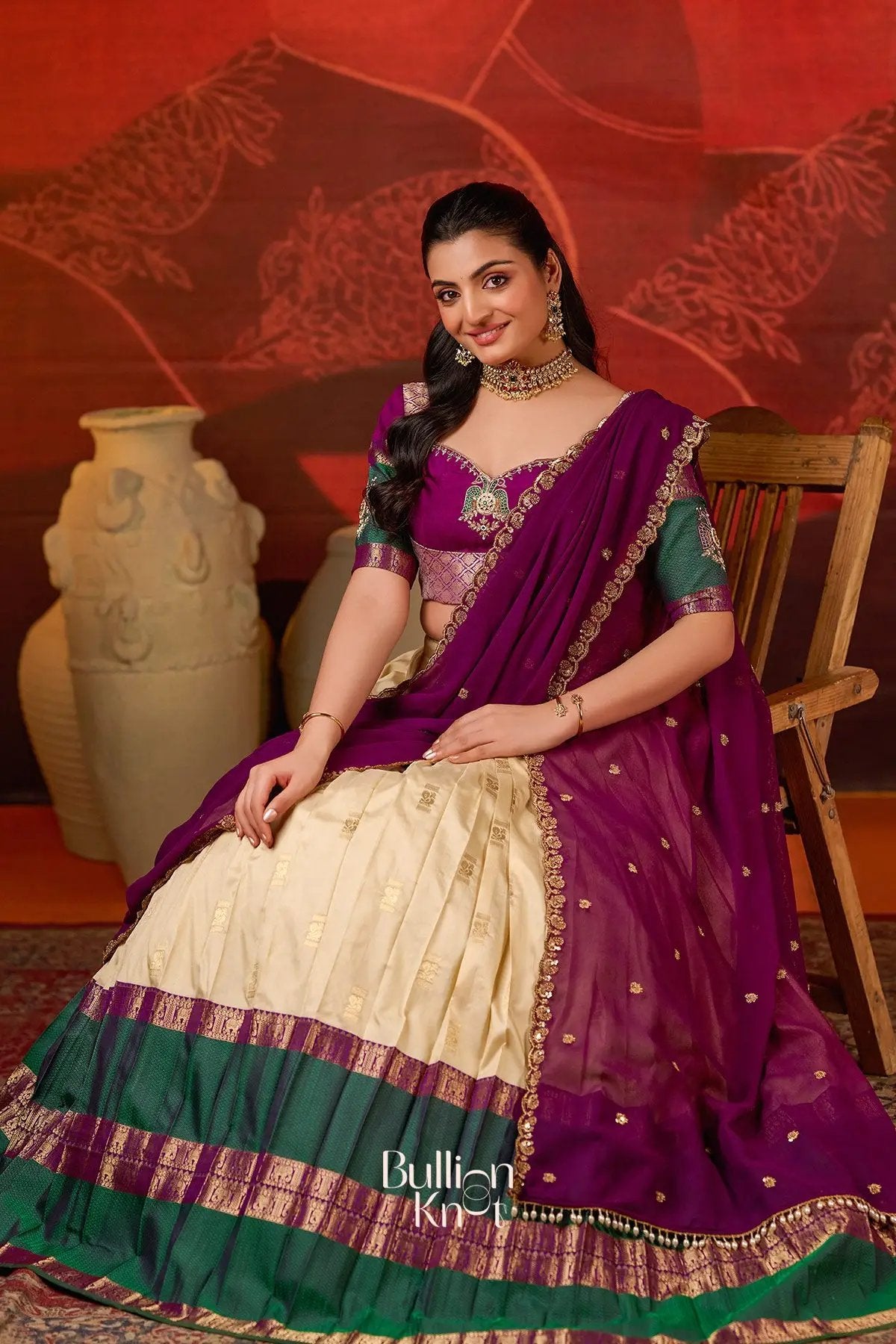 Model seated wearing Vishakha Half Saree in Vanilla at bullionknot, displaying the elegant vanilla skirt and rich purple dupatta with gold embellishments.