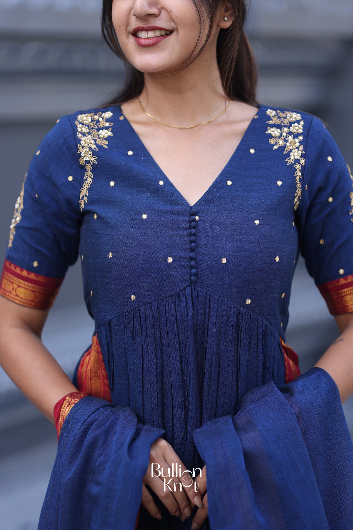 Woman wearing a blue traditional outfit with gold embroidery, posing against a blurred background.