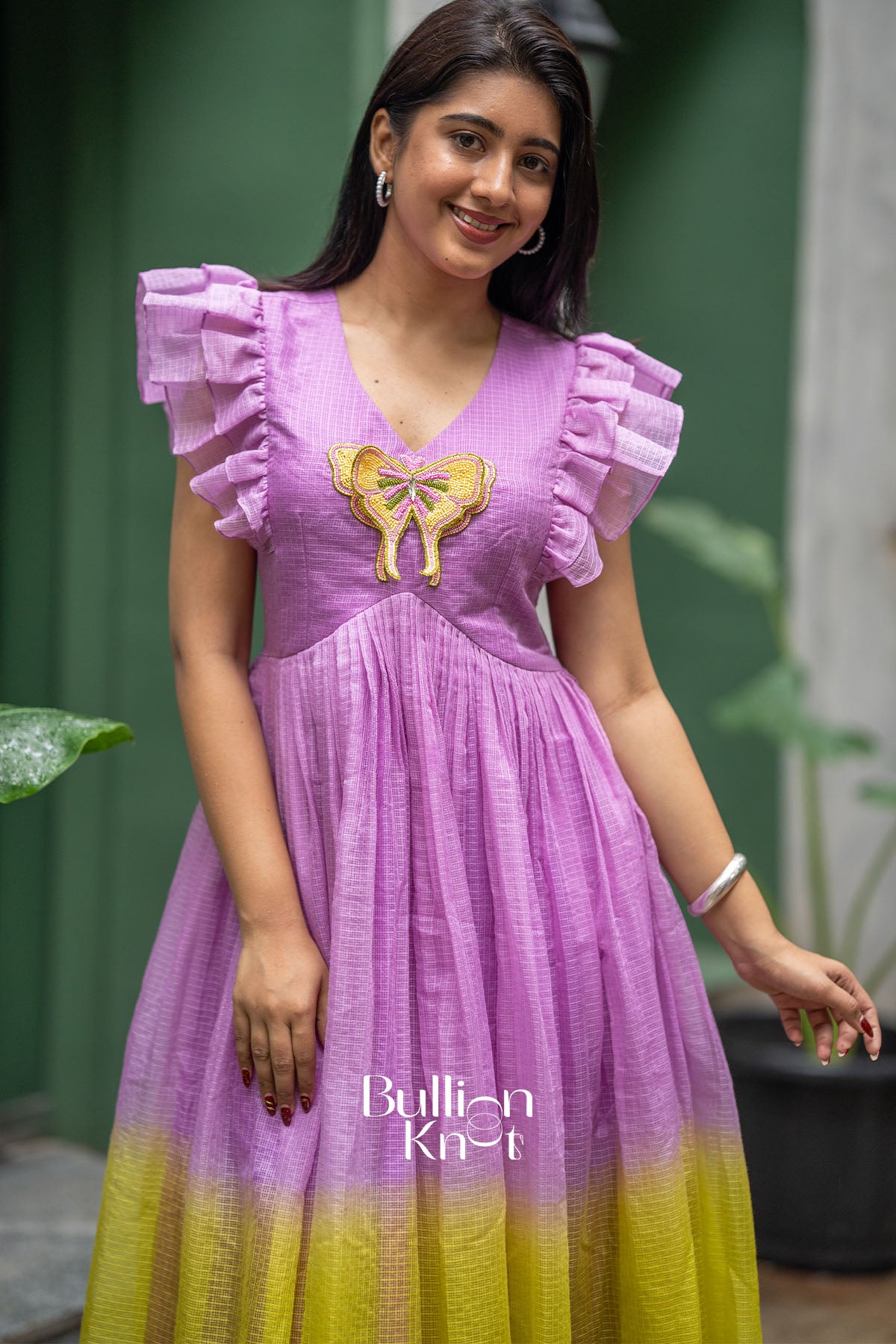 Woman wearing a purple and yellow dress with ruffled sleeves and a bow detail, standing outdoors.