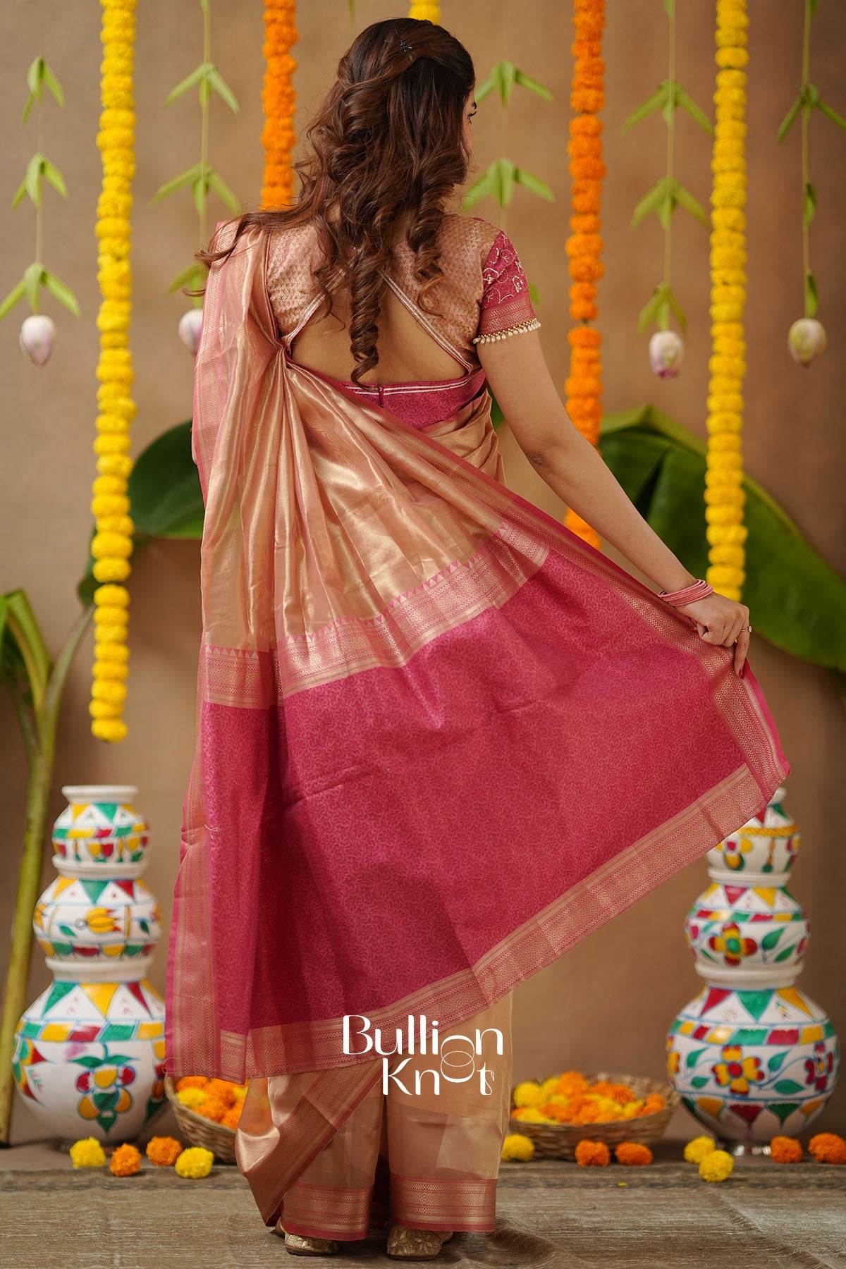 Woman in a pink saree with a decorative background featuring colorful pots and flowers.