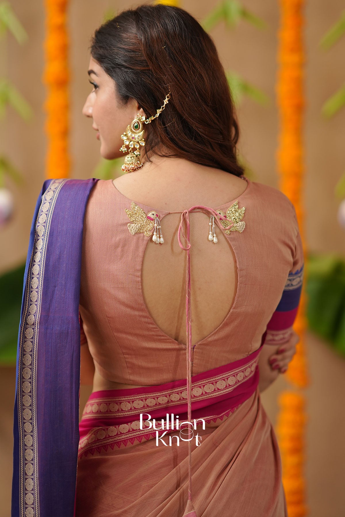 Woman wearing a traditional saree with a decorative back design, standing against a decorated background.