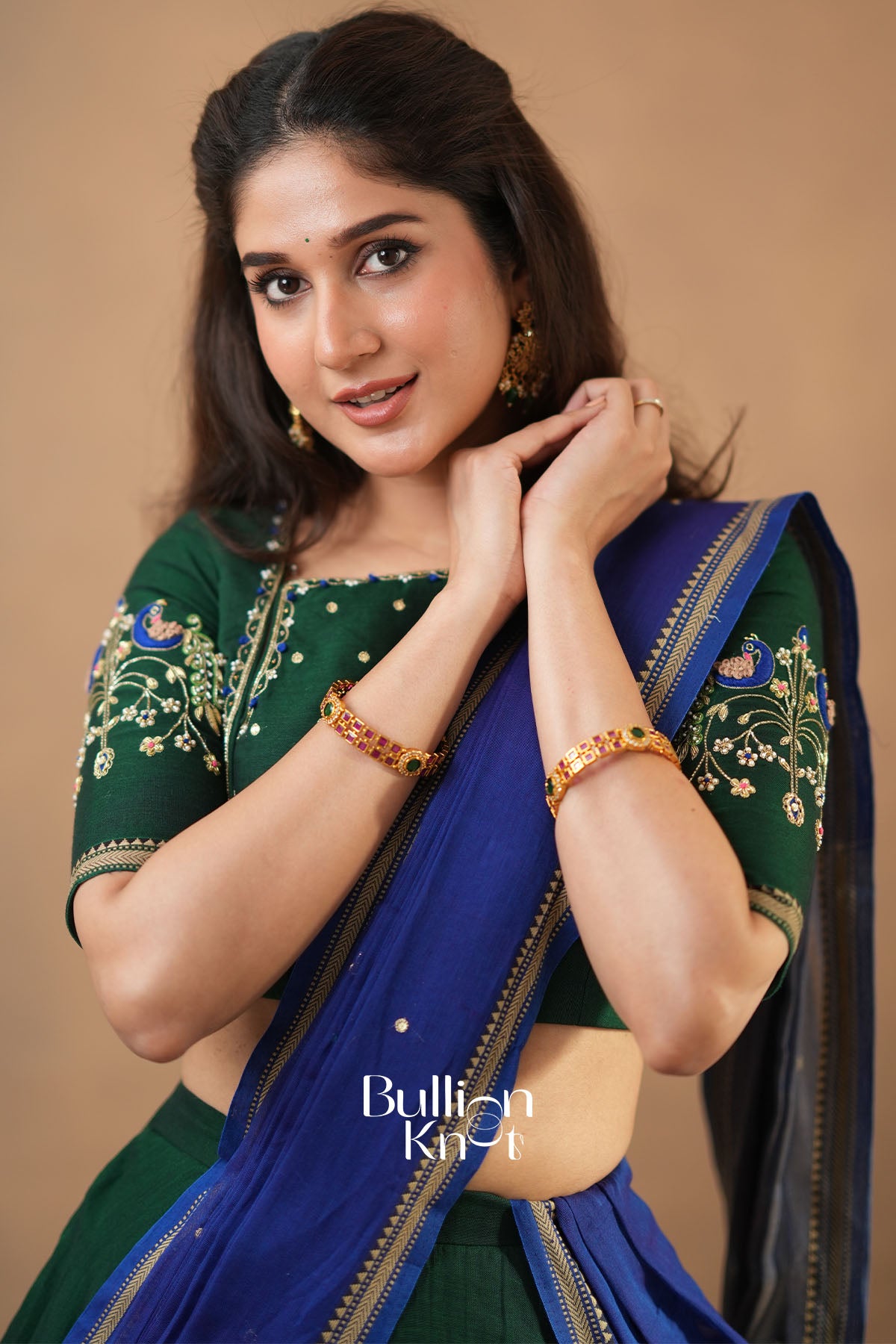 Woman wearing a green and blue saree with gold jewelry on a beige background