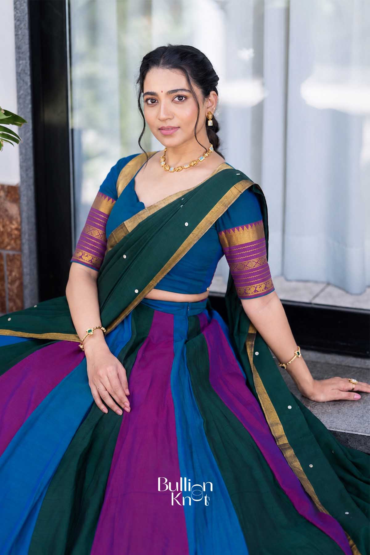 Woman sitting on stairs in a multicolor handloom half saree, showing back design with tie-up blouse and gold border, perfect for festive events.