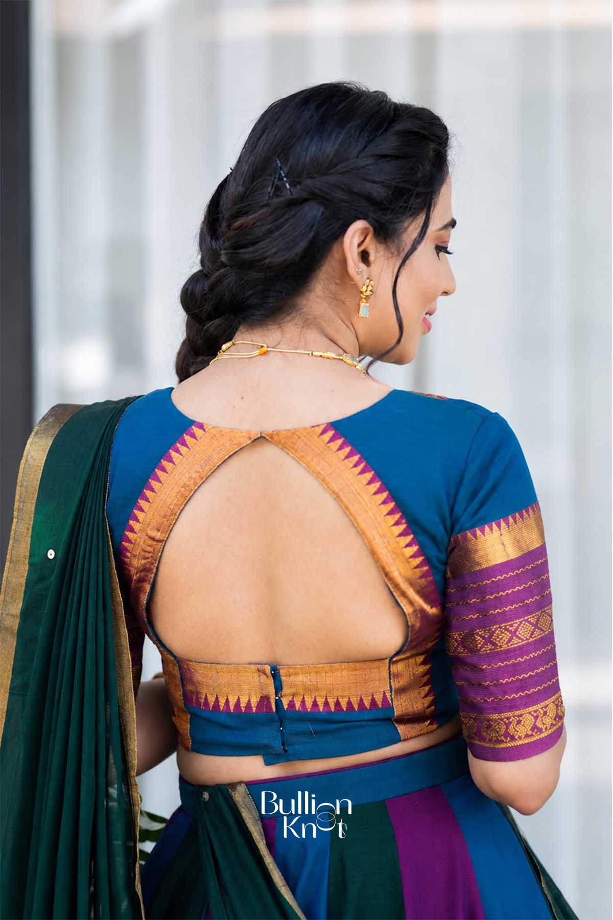 Side profile of woman in blue and purple saree, with braided hairstyle, gold jewelry, and elegant pose highlighting traditional festive wear.