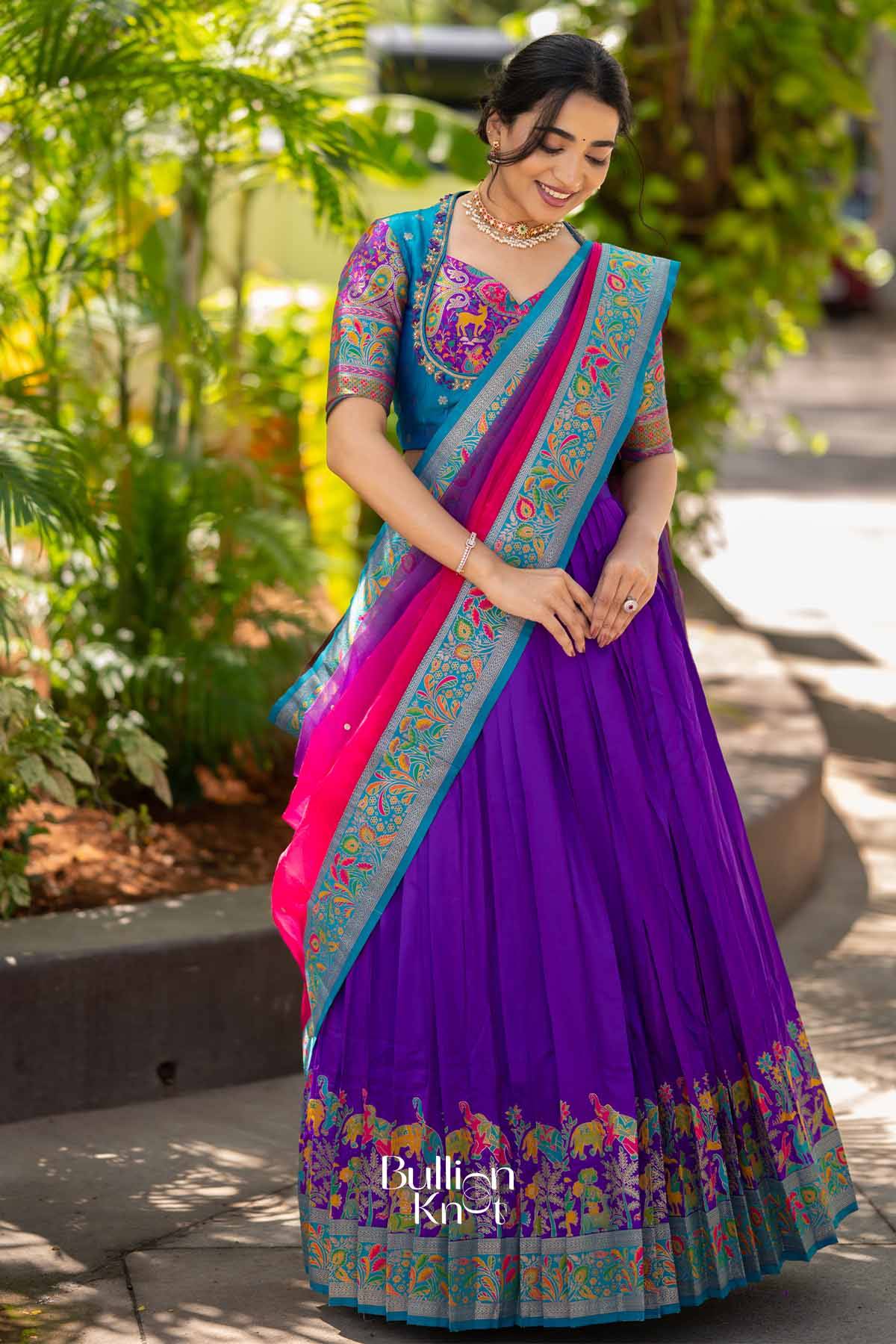 Woman in vibrant blue blouse with colorful embroidery, purple lehenga with embroidered border, smiling and adjusting earrings outdoors.