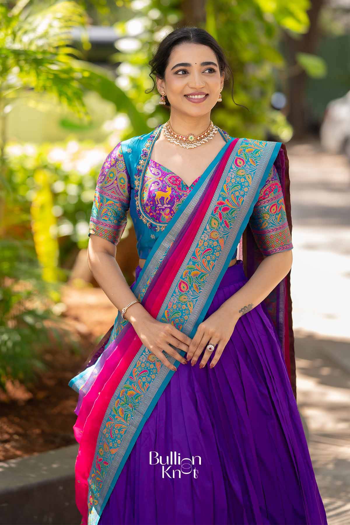 Front view of woman in purple silk lehenga with multicolored embroidered border, wearing traditional jewelry, smiling outdoors.