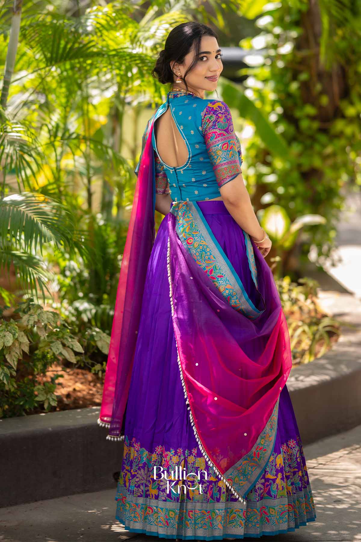 Back view of woman in blue blouse and purple lehenga, showcasing embroidered back design and matching dupatta with colorful border.