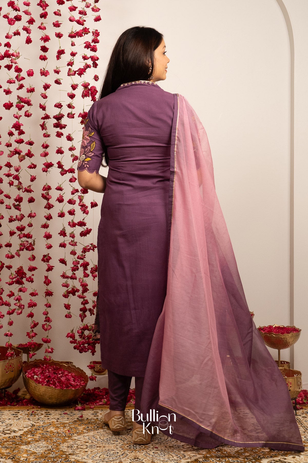 Back view of woman in purple Kalamkari suit with pink dupatta, floral wall decor