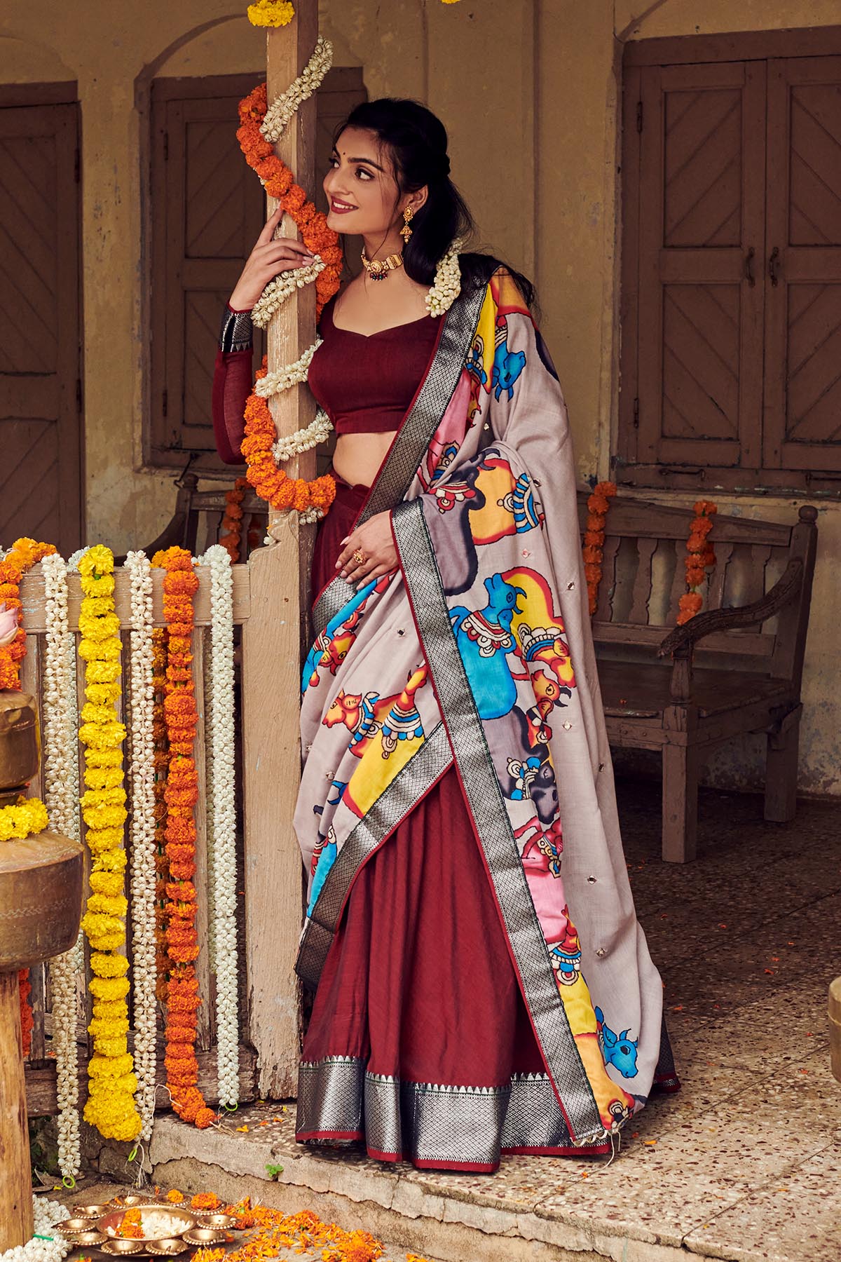 a women in dark maroon handloom lehenga dress with cotton dupatta playing with flowers at marriage function, dress name Shisha kalamkari Lehenga Set from Bullionknot