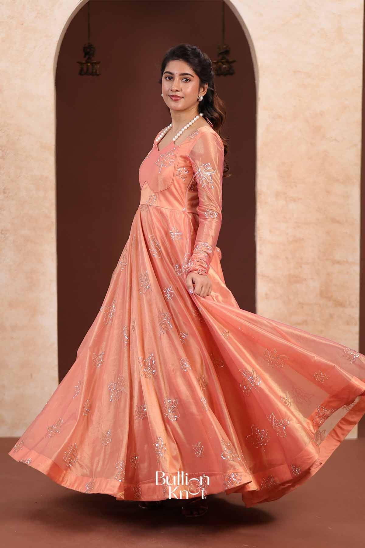 Elegant peach-colored Indo Western Anarkali gown with embroidered floral patterns, long sleeves, flowing skirt, and satin finish, perfect for special occasions.