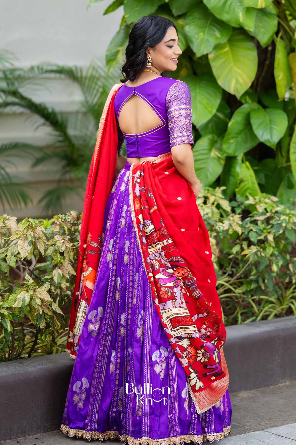 Back view of woman in purple blouse and red lehenga, showing intricate embroidery and open back design, outdoors