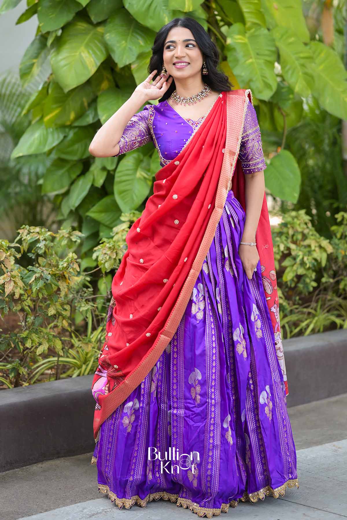 Elegant woman in purple and red traditional lehenga with intricate embroidery, jewelry, and lush green background