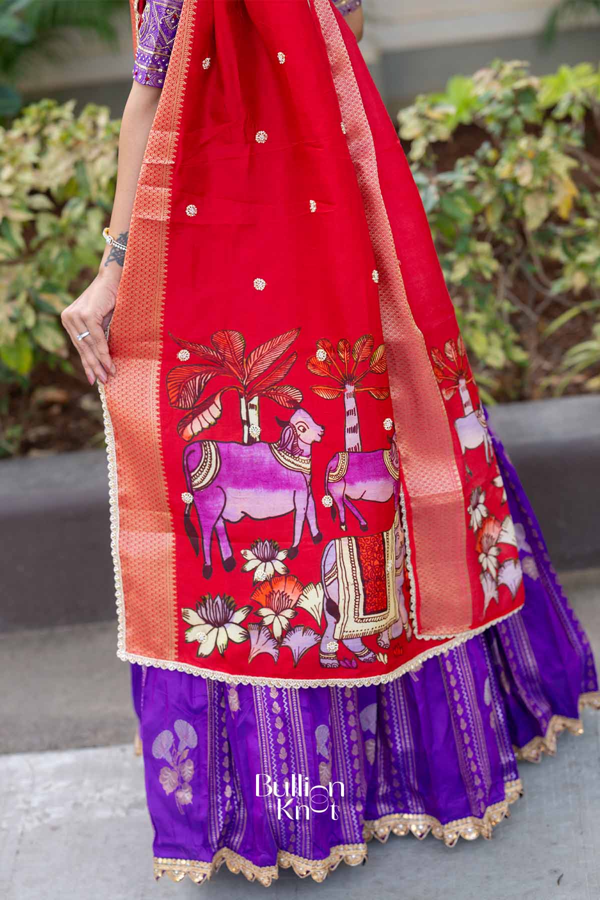 Close-up of woman’s hand holding red dupatta with floral and animal motifs, wearing jewelry, outdoors setting