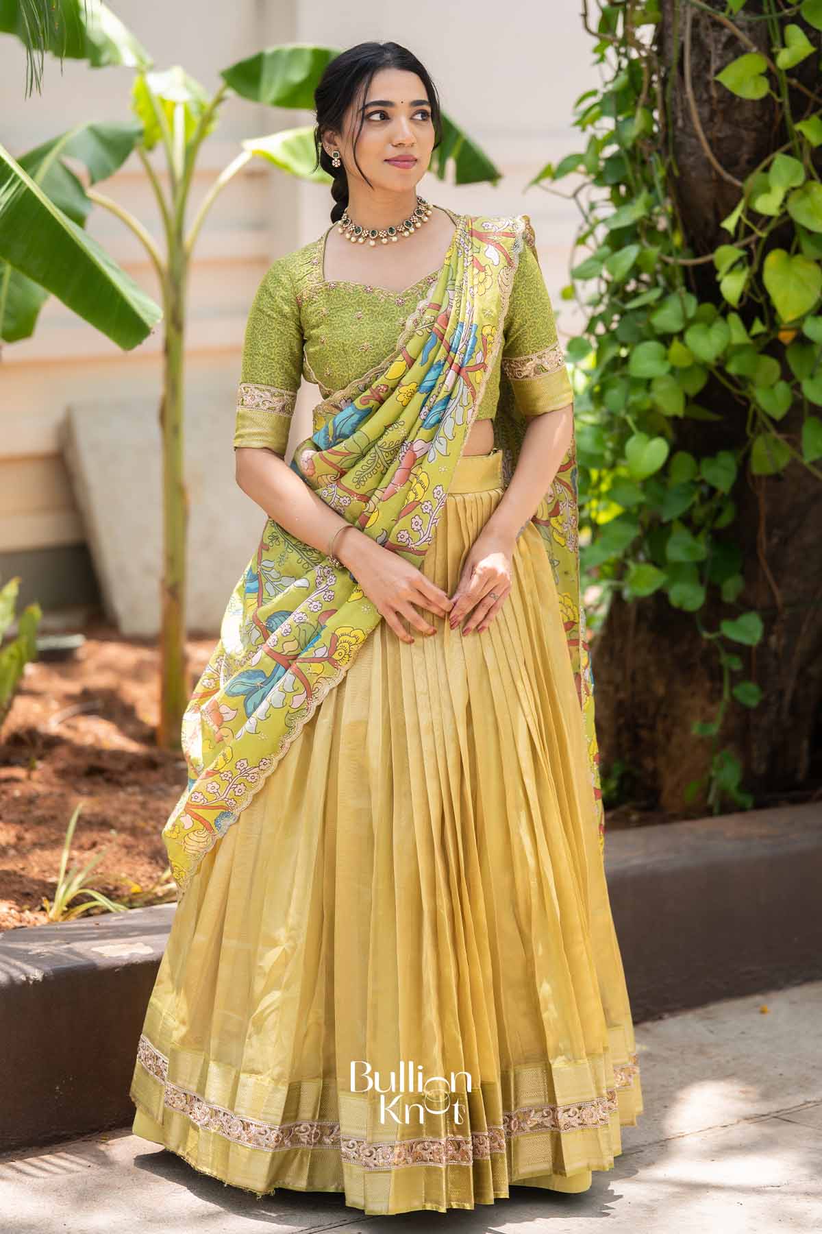 Elegant woman in yellow pleated lehenga with green embroidered blouse and floral dupatta, accessorized with jewelry, standing outdoors