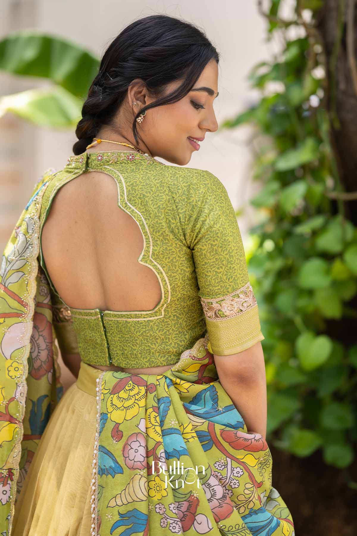 Side profile of woman smiling in yellow lehenga with floral dupatta, green embroidered blouse, and jewelry, outdoors with lush greenery.