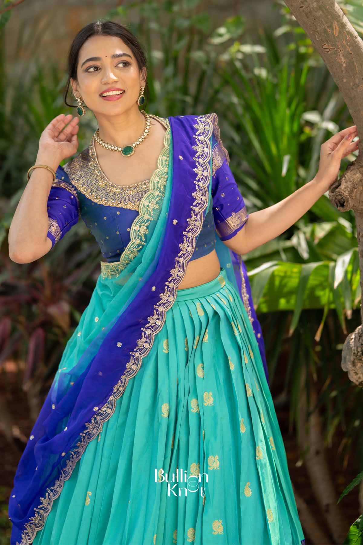 Close-up of woman in blue silk lehenga with intricate gold embroidery, wearing matching jewelry, smiling outdoors