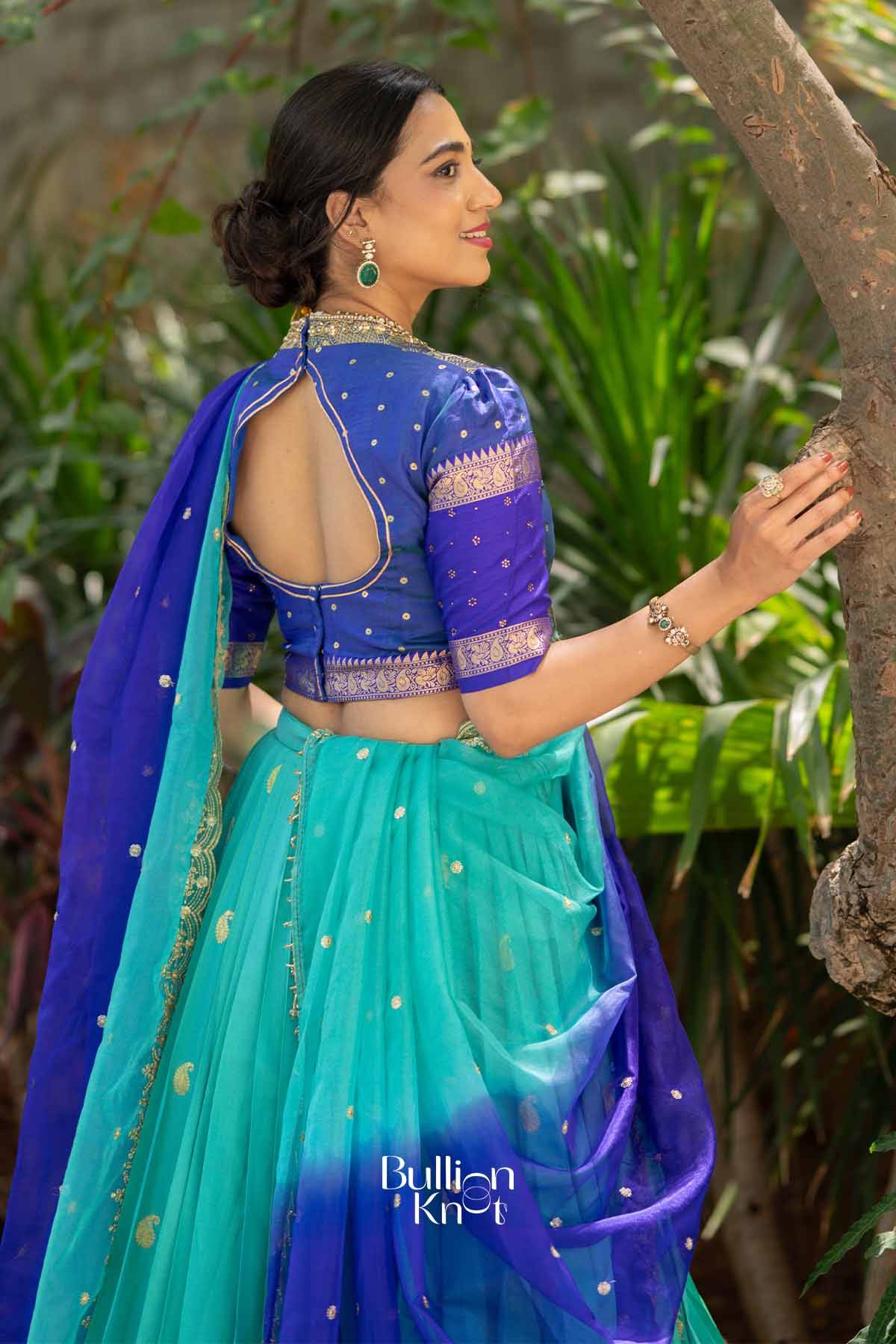 Elegant woman in blue silk lehenga with gold embroidery, open back blouse, matching jewelry, standing outdoors with greenery