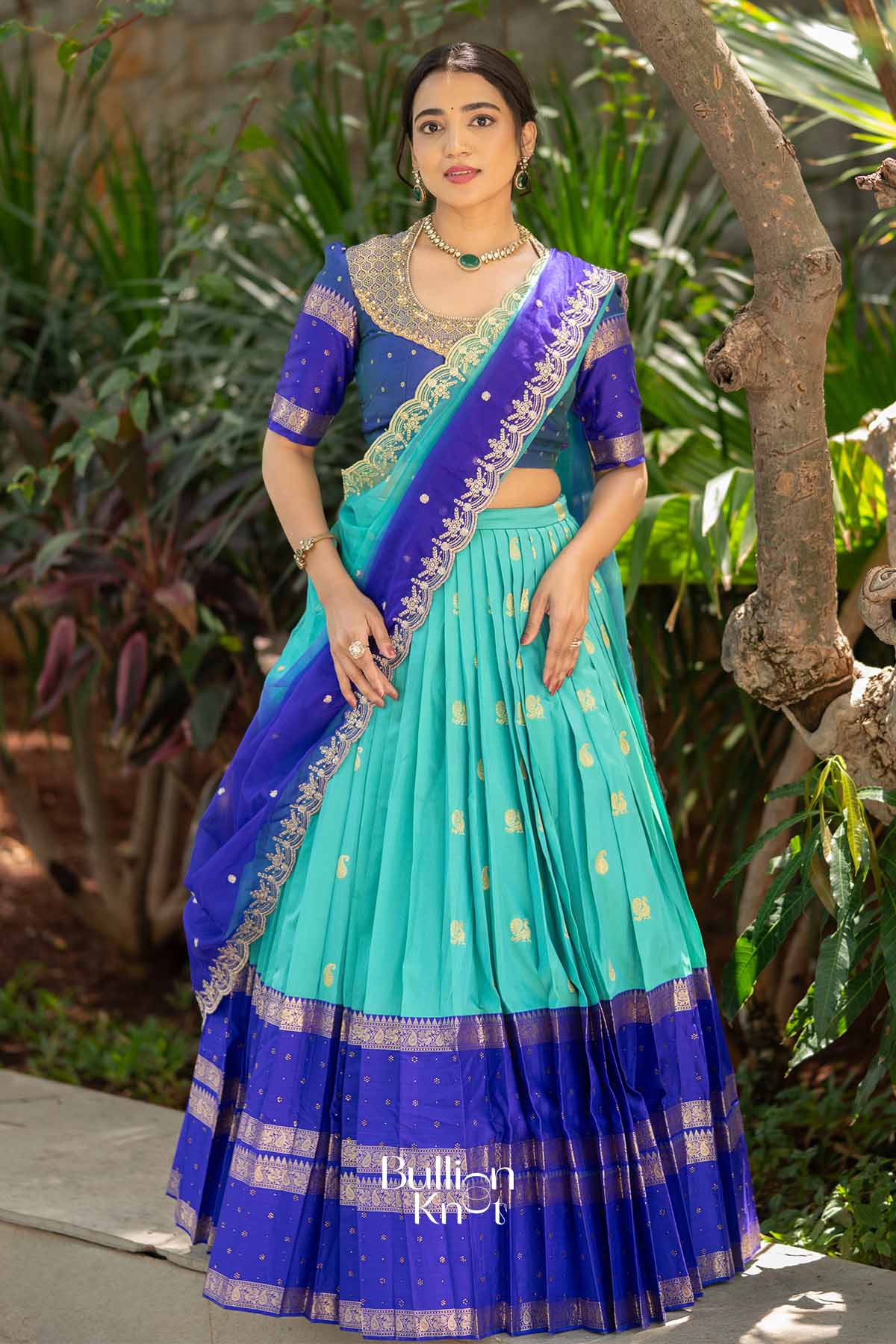 Portrait of woman in blue and turquoise lehenga with gold embroidery, jewelry, and greenery background